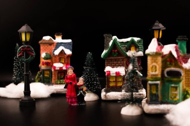 Karla kaplı bir ev ve ağaçların yanında şarkı söyleyen Noel şarkıları söyleyen minyatür Noel köyü sahnesi. Ilık, parlak pencereler, yumuşak odak ve karanlık arka plan nostaljik bir tatil atmosferi yaratır..