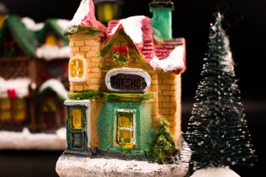 Karla kaplı bir ev ve ağaçların yanında şarkı söyleyen Noel şarkıları söyleyen minyatür Noel köyü sahnesi. Ilık, parlak pencereler, yumuşak odak ve karanlık arka plan nostaljik bir tatil atmosferi yaratır..