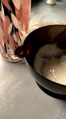 Siyah bir kâsede saç boyası karışımı ve profesyonel boya fırçası. Yumuşak ışık, koyu arkaplan ve kurutulmuş pampa otları ile kremsi bej rengi, sakin bir salon atmosferi yaratıyor..