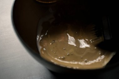 Siyah bir kâsede saç boyası karışımı ve profesyonel boya fırçası. Yumuşak ışık, koyu arkaplan ve kurutulmuş pampa otları ile kremsi bej rengi, sakin bir salon atmosferi yaratıyor..