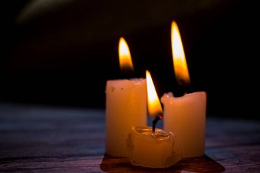 Üçgen şeklinde dizilmiş yanan mumların yakın çekimi. Koyu bir zemin, yumuşak bir alan derinliği ve şeffaf bir balmumu ile parlak alevler kontrastı samimi, atmosferik bir sahne yaratıyor..