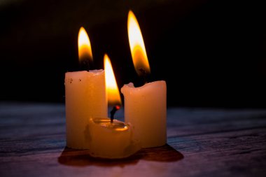 Üçgen şeklinde dizilmiş yanan mumların yakın çekimi. Koyu bir zemin, yumuşak bir alan derinliği ve şeffaf bir balmumu ile parlak alevler kontrastı samimi, atmosferik bir sahne yaratıyor..