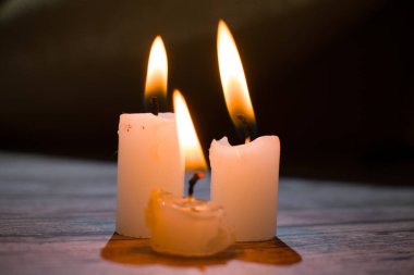 Üçgen şeklinde dizilmiş yanan mumların yakın çekimi. Koyu bir zemin, yumuşak bir alan derinliği ve şeffaf bir balmumu ile parlak alevler kontrastı samimi, atmosferik bir sahne yaratıyor..