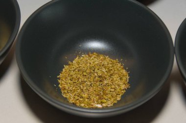 Yakından bakınca, beyaz bir yüzey üzerinde koyu gri bir seramik kasenin dibinde kurumuş kekik yaprakları görünüyor..