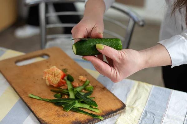 Kız bir bıçakla salatalık soyuyor. Beyaz bir masa örtüsünün arka planına karşı. Seçici odak.