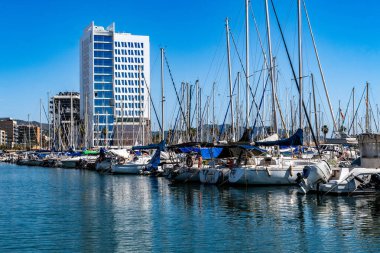 Nautical summer scene. View of moored yachts and sailboats in a port with a modern white high-rise building in the background. Concept of tourism, luxury travel and sailing lifestyle. Badalona, Spain