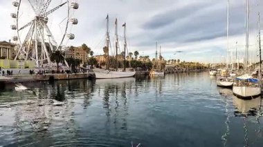 Barselona, İspanya-24 Kasım 2025. Turistler marinada tahta bir tahta kaldırımda büyük bir dönme dolap ve arka planda bir yelkenliyle yürüyorlar. Port Vell de Barcelona İspanya