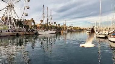 Barselona, İspanya-24 Kasım 2025. Turistler marinada tahta bir tahta kaldırımda büyük bir dönme dolap ve arka planda bir yelkenliyle yürüyorlar. Port Vell de Barcelona İspanya