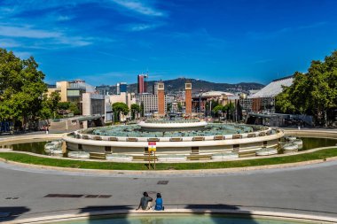 Barcelona 'daki Plaa d' Espanya 'ya doğru Montjuc Sihirli Çeşmesi' nin (Font Mgica) büyük dairesel havzasına panoramik bir bakış açısı. Arka planda ikonik Venedik kuleleri, şehir silueti ve Tibidabo dağı yer almaktadır.