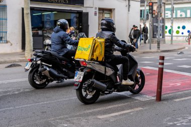 Barcelona, Spain-November 23, 2025. Two delivery riders on scooters in an urban setting, one with a yellow Glovo delivery bag, traffic signals and pedestrians in the background
