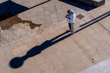 Uzun bir sabah gölgesi olan beton bir meydanda duran birinin yüksek açılı görüntüsü..