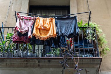Renkli giysiler, bir Barselona mahallesindeki yeşil bitkilerle dolu dövülmüş demir bir balkonun üzerinde asılı duruyor..