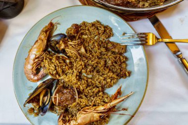 Yüksek açılı çekimde, çatal ve bıçağın yanında midye ve karidesli bir tabak deniz ürünleri pilavı görülüyor..