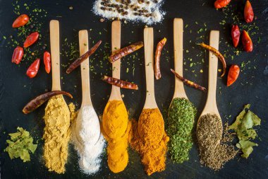 Çeşitli rengarenk baharatlar ve kurutulmuş otlar siyah tahta üzerinde kaşıklarda dizilmiştir..