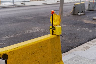 Üzerinde yanıp sönen bir uyarı işareti olan sarı bir trafik bariyeri inşaat ve yenileme çalışmaları yapılan bir caddede duruyor..