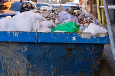 Büyük bir mavi metal çöp konteyneri yenileme sırasında bir şehir caddesinde enkaz ve plastik torbalarla dolup taşar..