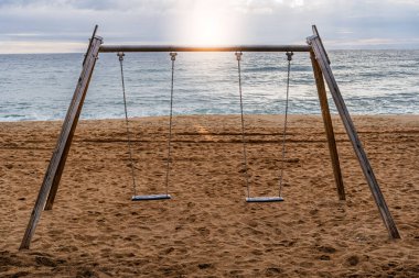 Denizin üzerinde güzel bir gün batımında kumlu bir sahilde sessizce sallanan iki salıncak. Kavram: Nostalji, sükunet ve günün sonu.