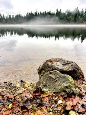 Sahil şeridi boyunca düşen yapraklarla, suların üzerinde yükselen büyük kayalar ve sabahın erken saatlerinde yüzeye yansıyan yemyeşil ağaçlar.