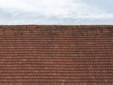 Bulutlu gökyüzünün önündeki eski çatı kiremitleri. Kırmızı çatı yıpranmış ve kirlenmiş. Üst üste binen kiremitler dış yüzeyin bir parçası olarak bir kalıp oluşturuyor..