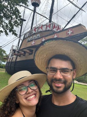 Genç, gülümseyen bir çift renkli tüyleri ve tarihi detayları olan etkileyici bir korsan gemisinin önünde poz veriyor. Etrafı yemyeşil, neşe ve macera dolu..