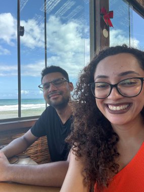 Gülümseyen bir çift, deniz kenarında büyük pencereli bir restoranda rahatlatıcı bir anın tadını çıkarıyor. Mavi gökyüzünün altında güzel okyanus manzarasını sergiliyorlar..