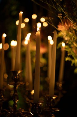 Ilık, parlak alevler ve arka planda yumuşak bokeh ışıklarıyla loş bir ortamda yanan uzun beyaz mumlar..