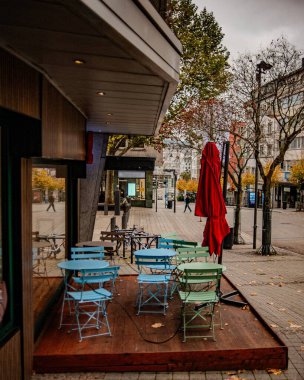 Katlanmış Şemsiyeli Renkli Kafe Terası, Pastel Tonlarda Boş Bistro Koltuklar, Yığılmış Metal Sandalyeler ve Küçük Masalar, Kırmızı Kapalı Örtü, Mağaza Önü,