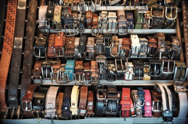 Bu canlı ve detaylı fotoğraf, muhtemelen bir zanaatkar pazarında ya da bir butik deri dükkanında el yapımı deri kemerlerin yoğun ve organize bir görüntüsünü yakalıyor. Kompozisyon, dokular, renkler ve malzemelerden oluşan zengin bir duvar halısıdır.