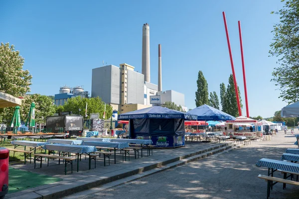 Pforzheim Almanya Juni 5, 2018 güneşli şehir bira bahçesi boş piknik masaları ve mavi şemsiyelerin altındaki ahşap banklar kompakt büfeleri, yüksek kuleli bacalar ve arka plandaki kırmızı direkler,