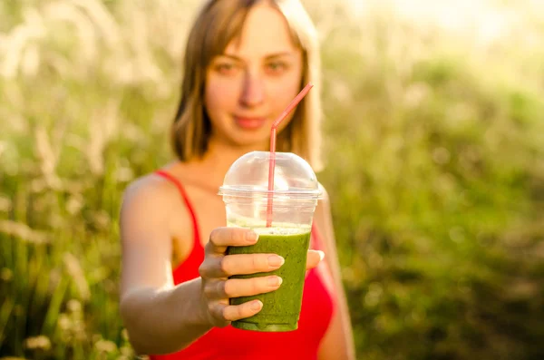 Лицо улыбающейся молодой женщины в руках зеленая энергия и питательный коктейль смузи на фоне размытой природы — стоковое фото