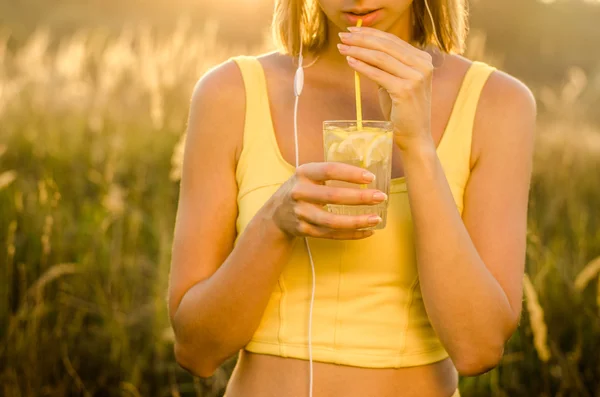 Healthy young girl drinker refreshing lemonade with lemon slices on a transparent glass cup high on blurred background nature — Stock Photo, Image