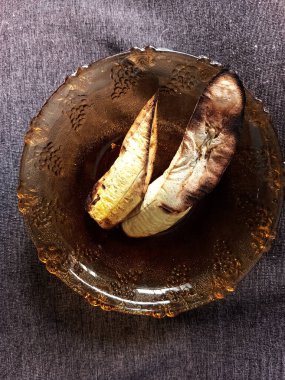 Kahverengi masa örtüsünün üzerindeki küçük sarı cam kasede kavrulmuş muz parçaları. Küçük boy ızgara muz. Kuru sıcak ızgara, evde pişirme metodu. Dilimlenmiş muz servisi.