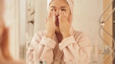 Beyaz bornoz ve kafasında havlu olan genç bir kadın evde masaj pedleri kullanarak cilt ürünleri, yüz maskeleri ve toner kullanıyor. Rahatlatıcı kişisel bakım ve güzellik rutin konsepti