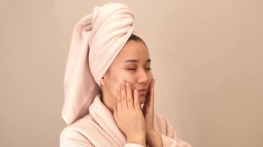 Beyaz bornoz ve kafasında havlu olan genç bir kadın evde masaj pedleri kullanarak cilt ürünleri, yüz maskeleri ve toner kullanıyor. Rahatlatıcı kişisel bakım ve güzellik rutin konsepti