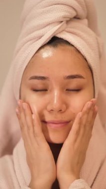 Beyaz bornoz ve kafasında havlu olan genç bir kadın evde masaj pedleri kullanarak cilt ürünleri, yüz maskeleri ve toner kullanıyor. Rahatlatıcı kişisel bakım ve güzellik rutin konsepti