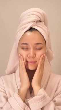 Beyaz bornoz ve kafasında havlu olan genç bir kadın evde masaj pedleri kullanarak cilt ürünleri, yüz maskeleri ve toner kullanıyor. Rahatlatıcı kişisel bakım ve güzellik rutin konsepti