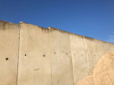 Mavi gökyüzünün önünde kum dağı olan beton bir duvar. Pürüzsüz, dalgalı bir kum tepeciğinin arkasında devasa beton duvarlı minimalist sanayi manzarası. İnsanların yokluğu geometrik basitliği vurgular, yalnızlığı ve dinginliği hatırlatır..