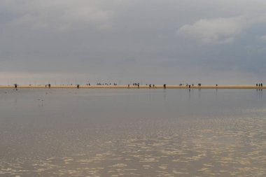 Sandbar where small figures stroll along the shore. Reflective water in the foreground mirrors the soft blues and grays of the overcast sky, creating a calm, muted atmosphere.