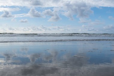 Ufukta dalgalar beliren sakin bir sahil. Yumuşak maviler ve gri renkler gökyüzü ve okyanusta karışıyor, ıslak kumda yansıyor, barışçıl bir hava yaratıyor. Küçük köpük bantlar kıyı şeridini işaretliyor, denizin ritmik hareketini işaret ediyor..