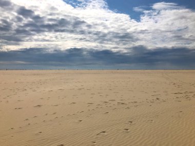 Uçsuz bucaksız kum ve bulutlarla dolu dramatik gökyüzü ile sakin kumlu bir sahil manzarası. Sahne, doğa meraklıları ve fotoğrafçılar için mükemmel bir huzur ve doğal güzellik hissi uyandırıyor..