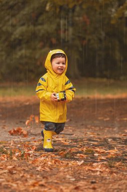 Parlak sarı yağmurluk ve uyumlu lastik botlar giyen mutlu genç bir çocuk, parkta ıslak bir yolda duruyor, gülümsüyor ve elinde bir oyuncakla yumuşak sonbahar yağmurunun tadını çıkarıyor.