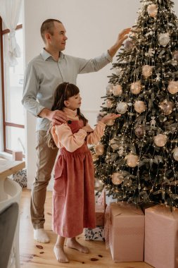Mutlu baba ve kız Noel ağacını parlak ışıklar ve süslemelerle donatarak, paketlenmiş hediyelerle çevrili, rahat, aydınlık, modern bir evde. Yeni yıl, Noel
