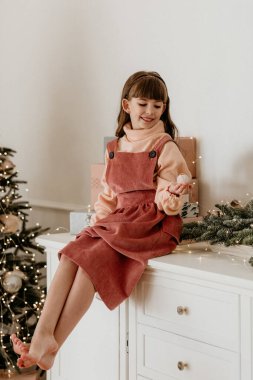 Mutlu çocuk bir şifonyerin üzerinde çıplak ayakla oturuyor, küçük bir süs taşıyor, hediye paketleriyle çevrili, ve rahat bir odada bulanık bir Noel ağacı.