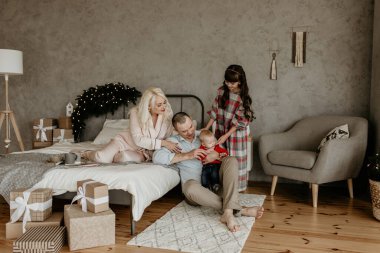 Dört kişilik mutlu bir aile Noel için dekore edilmiş şık ve modern bir yatak odasında bebekleriyle oynuyor ve etrafı hediyelerle çevrili.