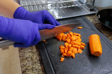 Elleri parlak mavi hijyenik eldivenlerle yakın plan çekimde mutfaktaki koyu renk bir kesim tahtasının üzerinde büyük bir şefin bıçağıyla çiğ havuçları küçük küplere ayırıyorlar. Yemek pişirmek. Tarifler