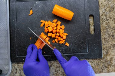 Mavi hijyenik eldivenlerle mutfak tezgahında plastik bir kesme tahtasının üzerinde çiğ havuçları küçük küplere doğrayan ve yemek hazırlayan el resimleri. Yemek pişirmek. Tarifler