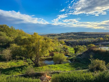 Yuvarlanan tepelerin ve vadilerin hareketli manzarası, küçük bir gölet, altın ve yeşil sonbahar yapraklarıyla kaplı, dramatik mavi gökyüzü altında dağınık beyaz ve gri bulutlar