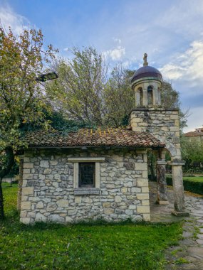 Küçük, kırsal bir Ortodoks taştan şapelin dikey görüntüsü terasotta kiremitli çatısı ve bakır kubbeli taş bir çan kulesi, bulutlu bir sonbahar gününde yeşil bir avluda geçiyor..
