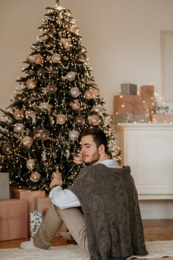 Dikiz manzaralı, kalın örgü şallı ya da pançolu genç bir adamın parlak ışıklandırılmış bir Noel ağacının yanında yere oturup kameraya bakarken çekilmiş bir portresi.