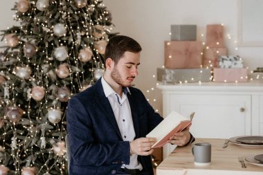 Genç, zarif giyimli bir adam bir masada oturuyor, kitap ya da günlük okuyor ve kahve içiyor, göz kamaştırıcı, sıcak bir Noel ağacı ve arka planda paketlenmiş hediyelerle.